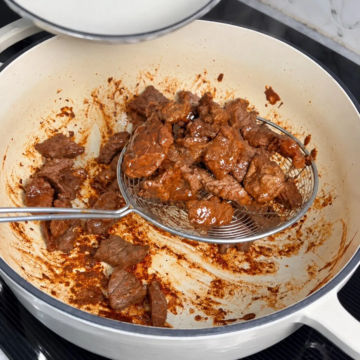 Browned beef pieces lifted from a Dutch oven with a wire skimmer.