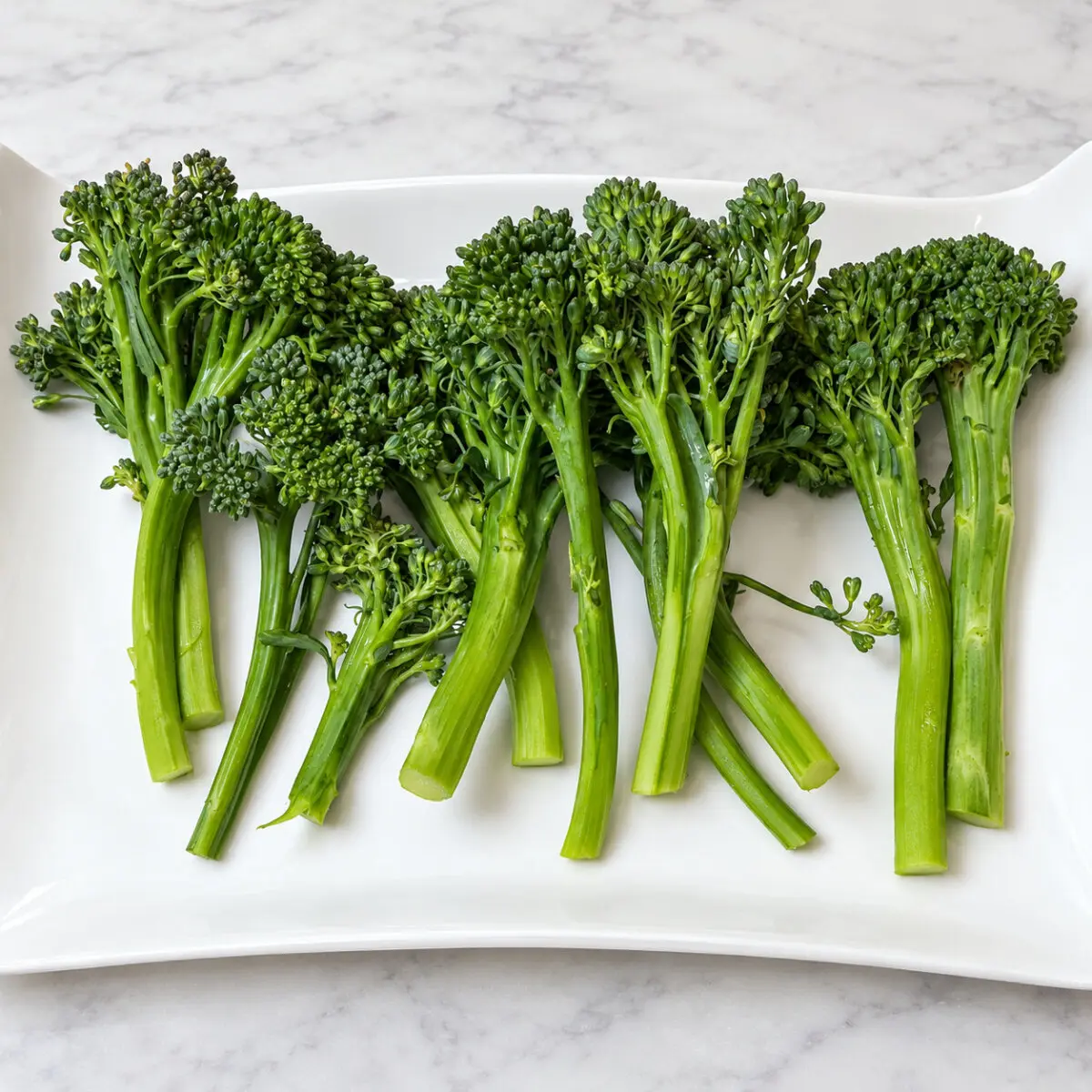 Broccolini stalks arranged on a white rectangular platter on a pale marble countertop.