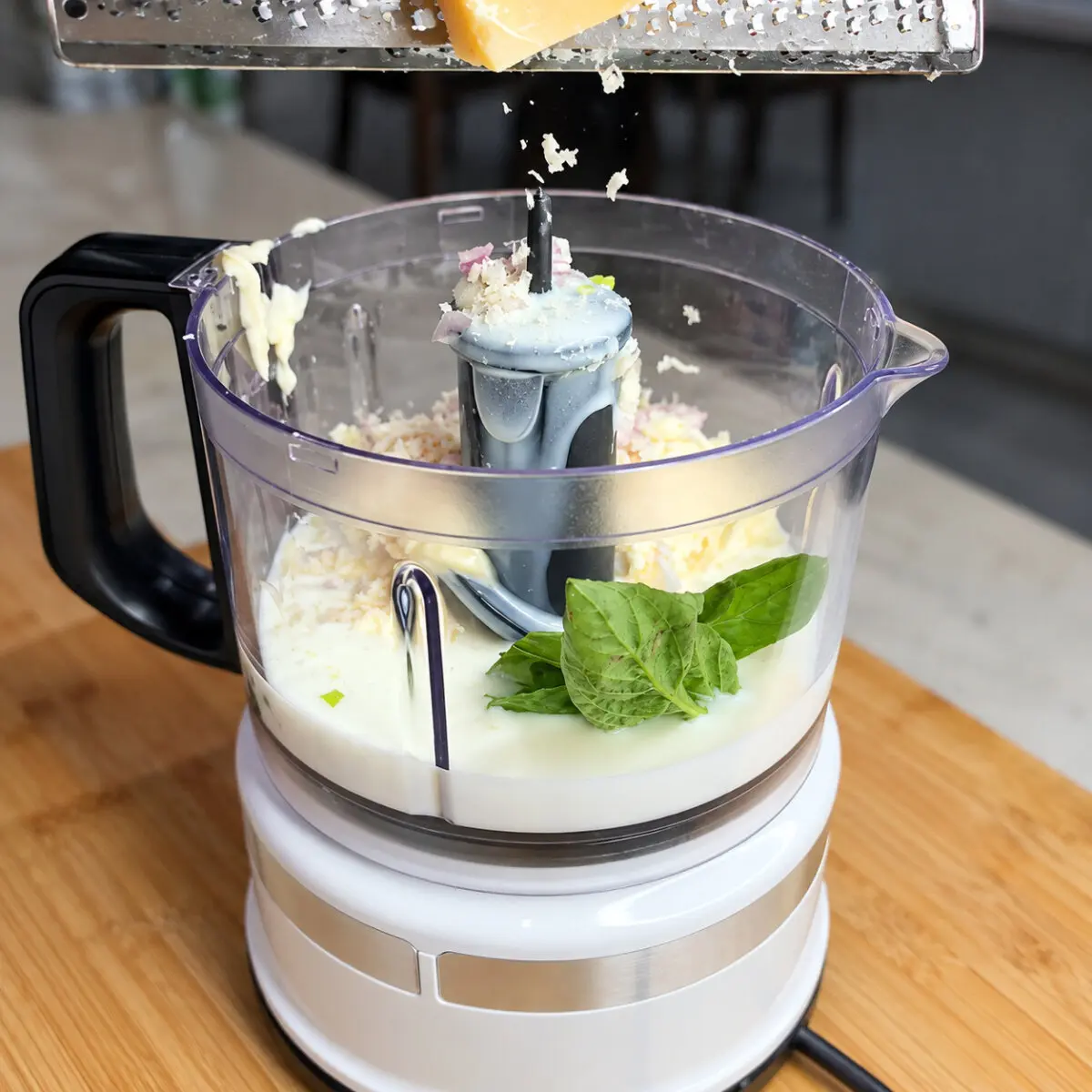 Parmesan being grated into a food processor containing creamy dressing and fresh basil.