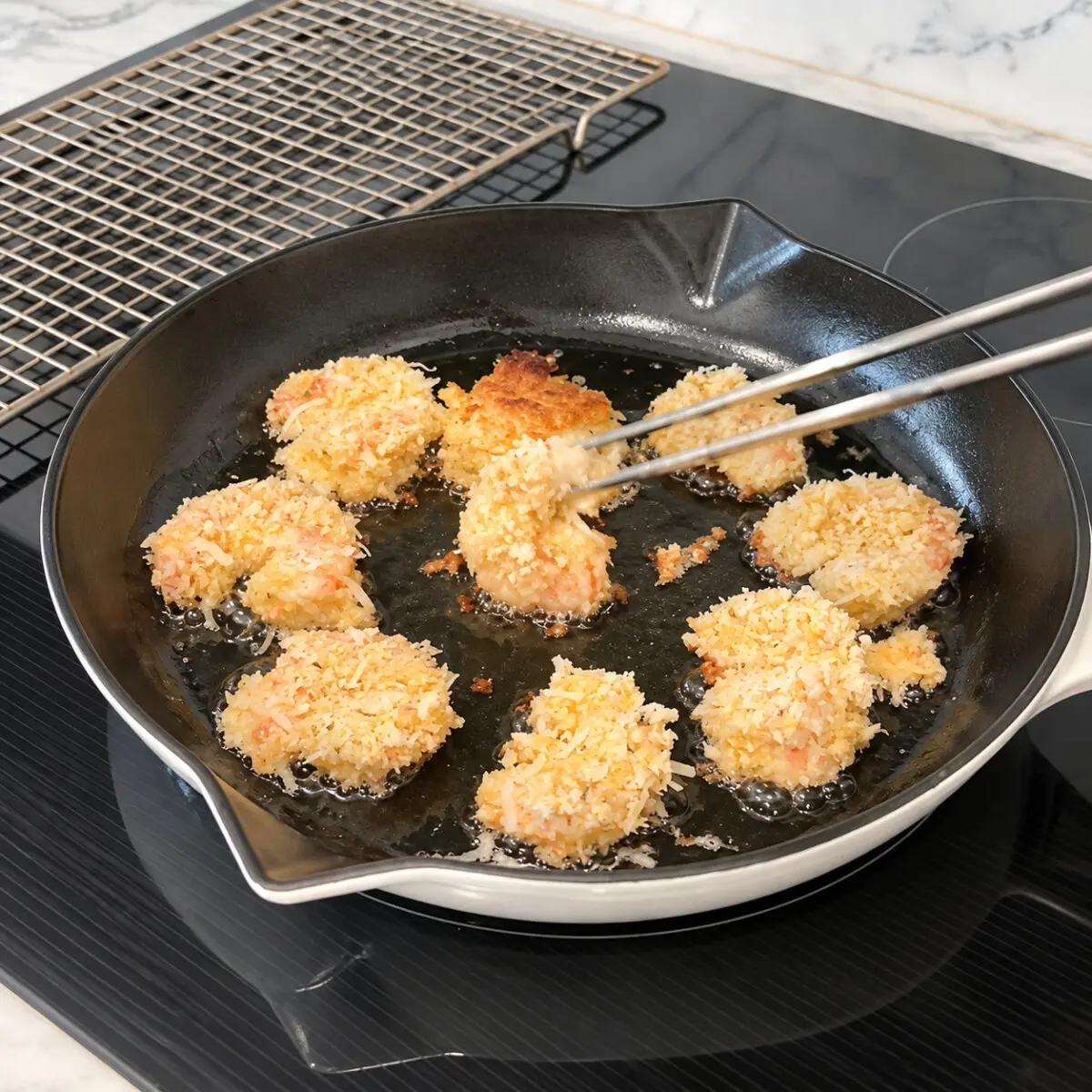 Coconut-coated shrimp frying in a black pan, with metal tongs turning a shrimp and a wire rack behind the pan.