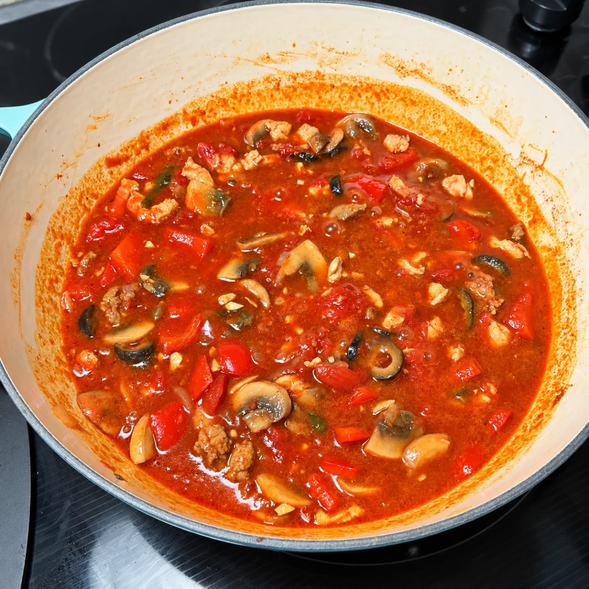 Chicken, mushrooms, peppers and olives simmering in tomato sauce in a turquoise pan.